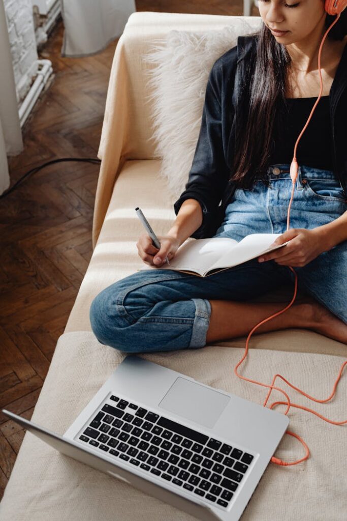 Young woman studying at home on a couch with a laptop and notebook, wearing headphones.